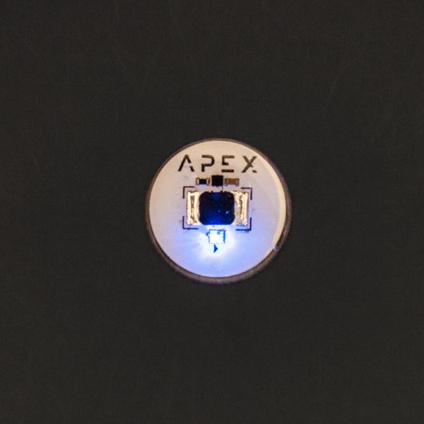 暗い背景に置かれたApex Spectrum。銅色の縁とAPEXの文字が入った円形のインプラント。青い光が点灯している